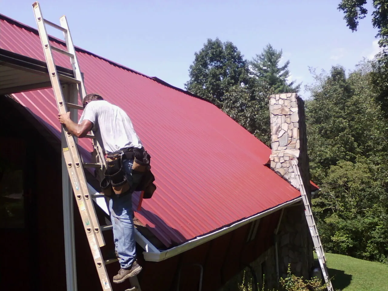 Roofer on a ladder beside a red metal roof and stone chimney — Soffit & Fascia Repair by Roofing Brothers in Muskego